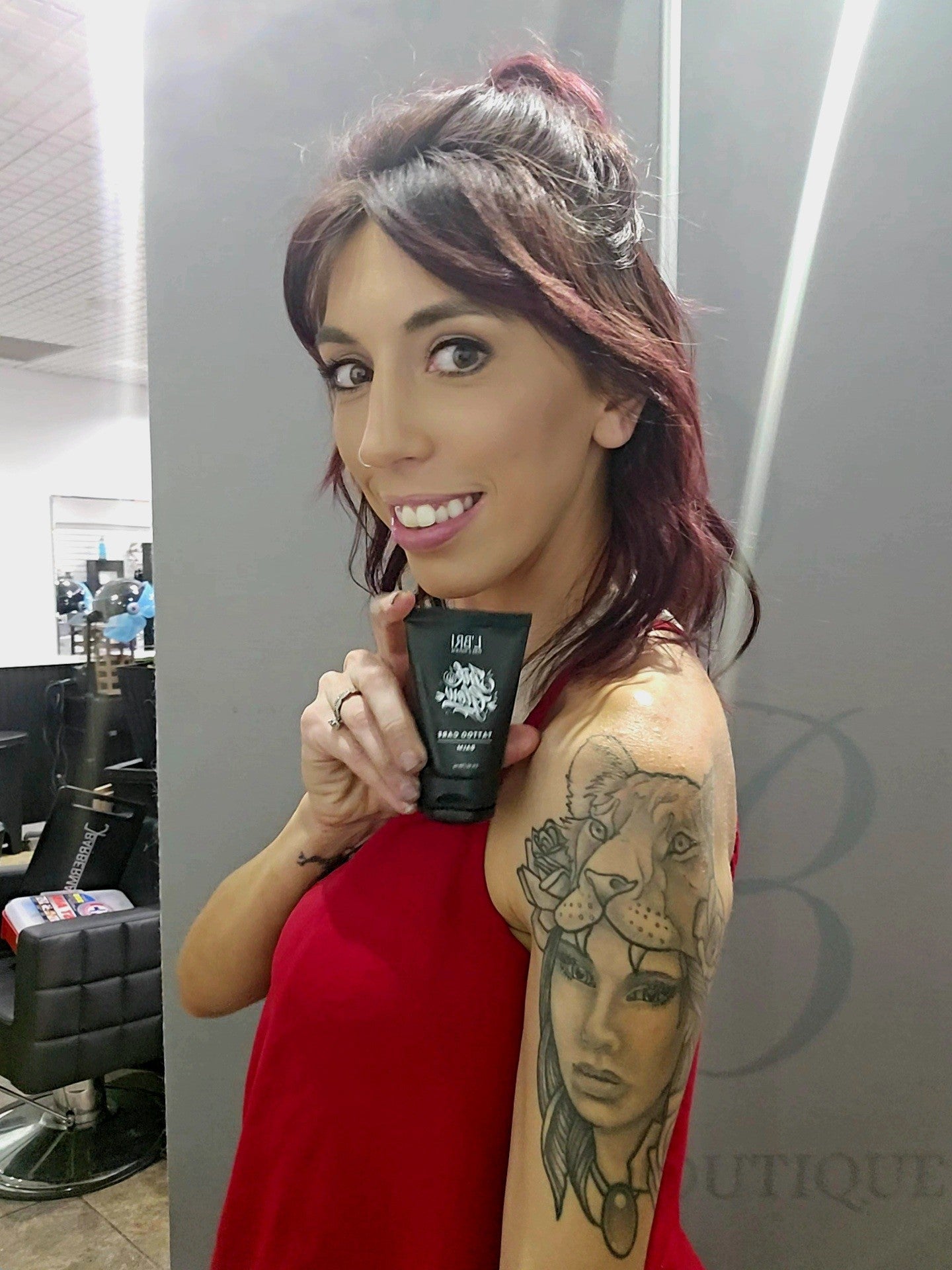 Woman holding a black tube of product with visible tattoos, standing in a salon.
