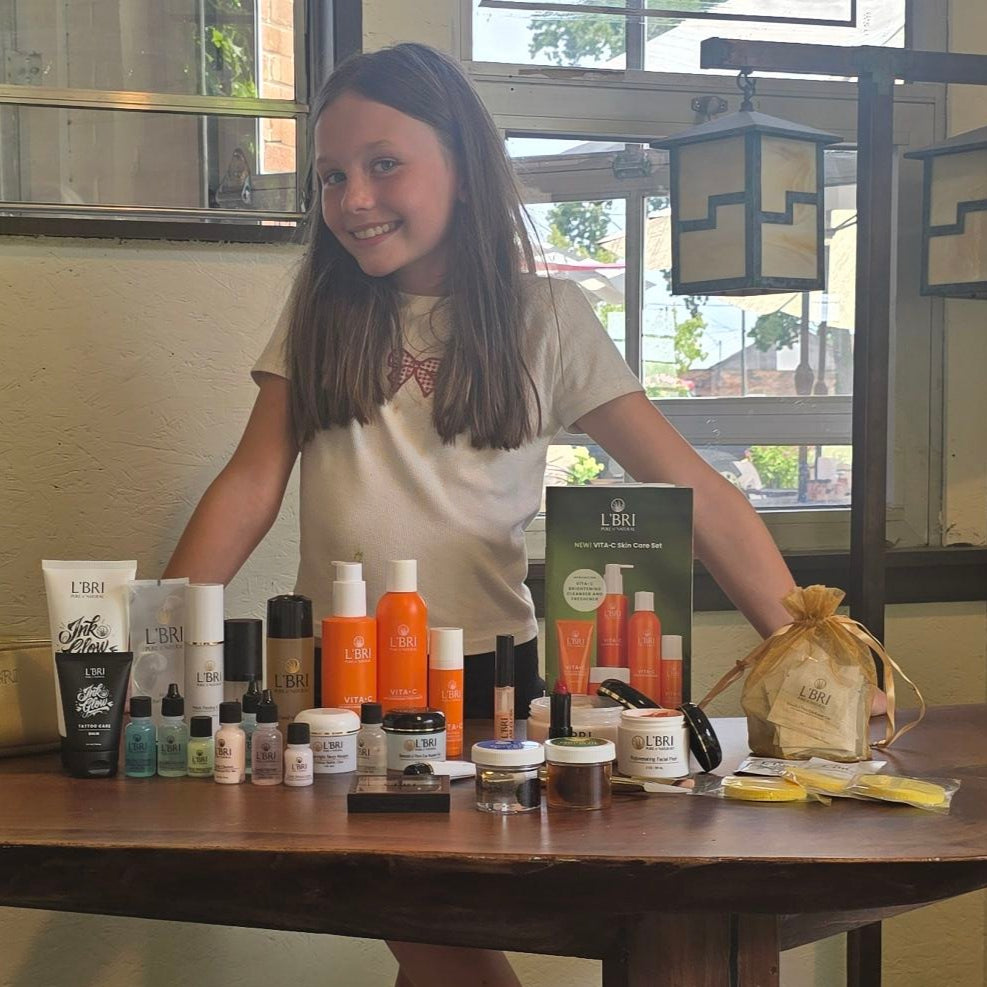 Young girl standing behind a table with various skincare products and a box labeled 'LBU' in a room with windows.