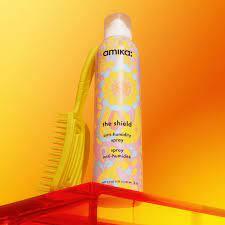 A bottle of Amika The Shield heat protection humidity blocker spray with a yellow label, displayed on a yellow background, accompanied by a yellow hairbrush.