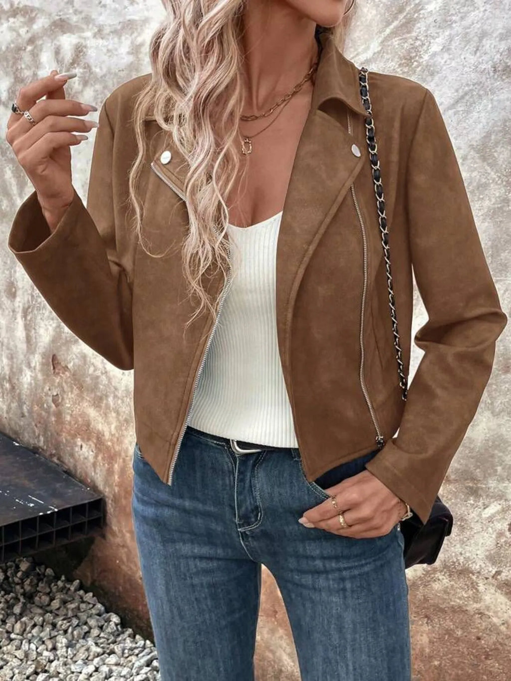 Person wearing a brown suede jacket, white top, and blue jeans against a textured wall.