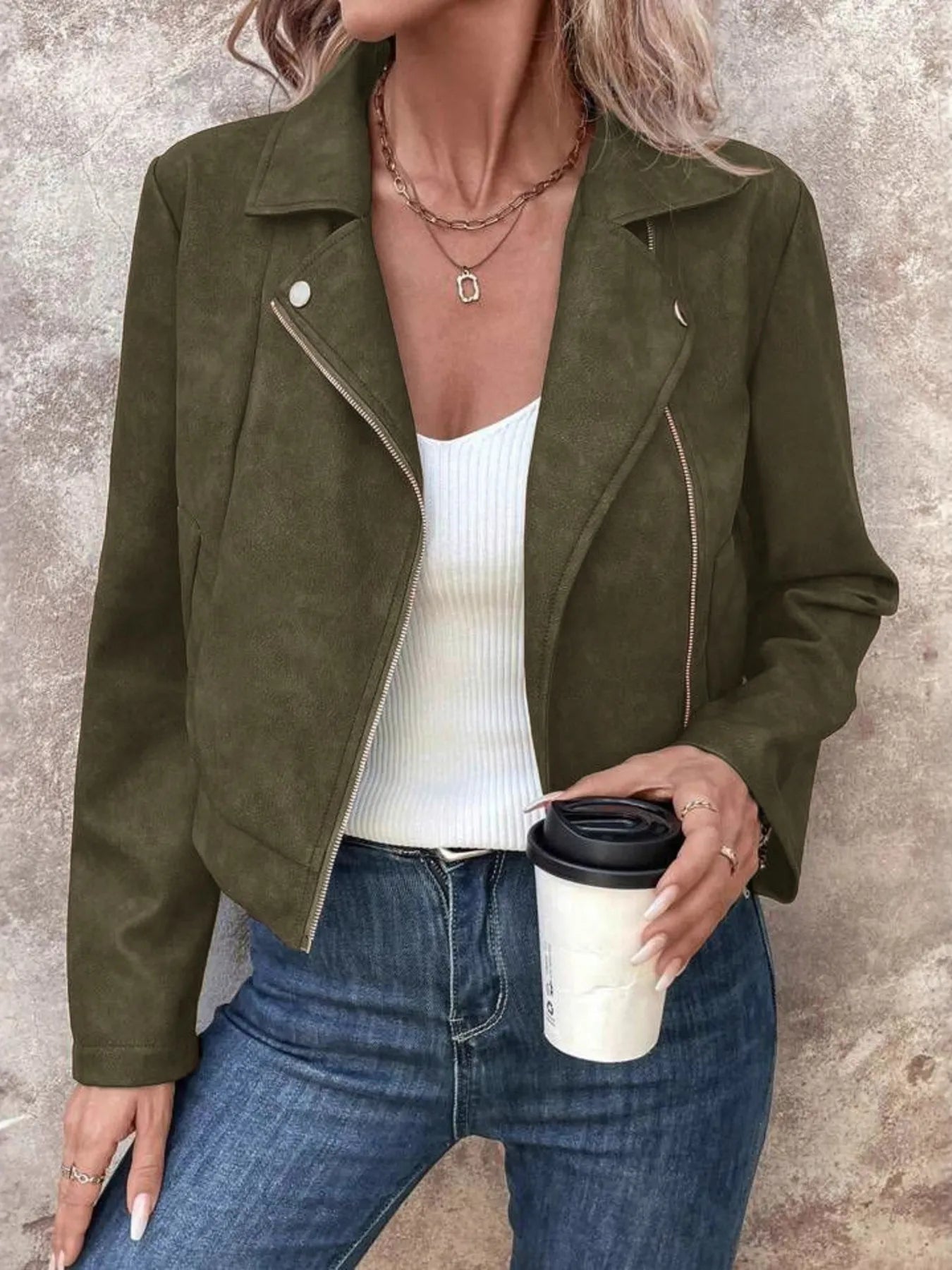 Person wearing a green suede jacket, white top, and blue jeans, holding a coffee cup.