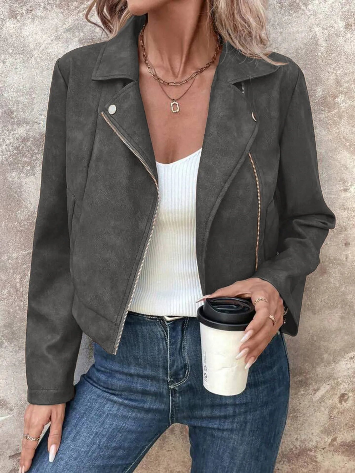 Person wearing a dark gray suede jacket, white top, and blue jeans, holding a coffee cup.