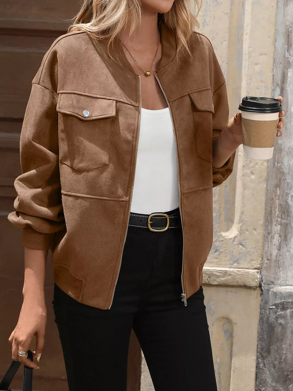 Person wearing a brown jacket holding a coffee cup against a textured wall.