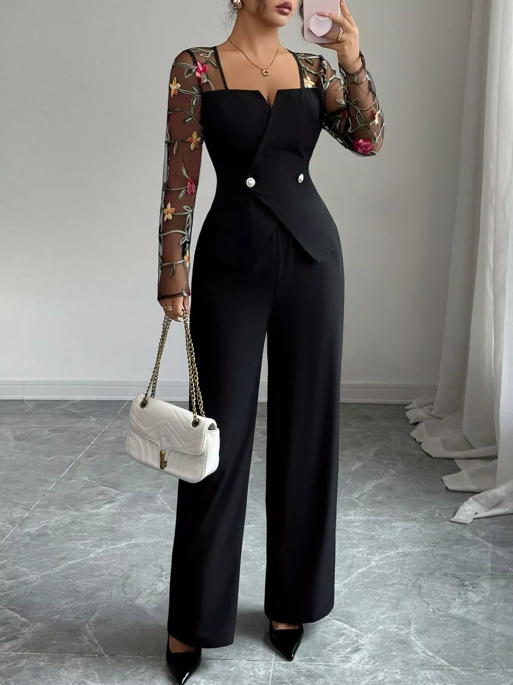 Person wearing a black outfit with floral sleeves, holding a white handbag indoors.