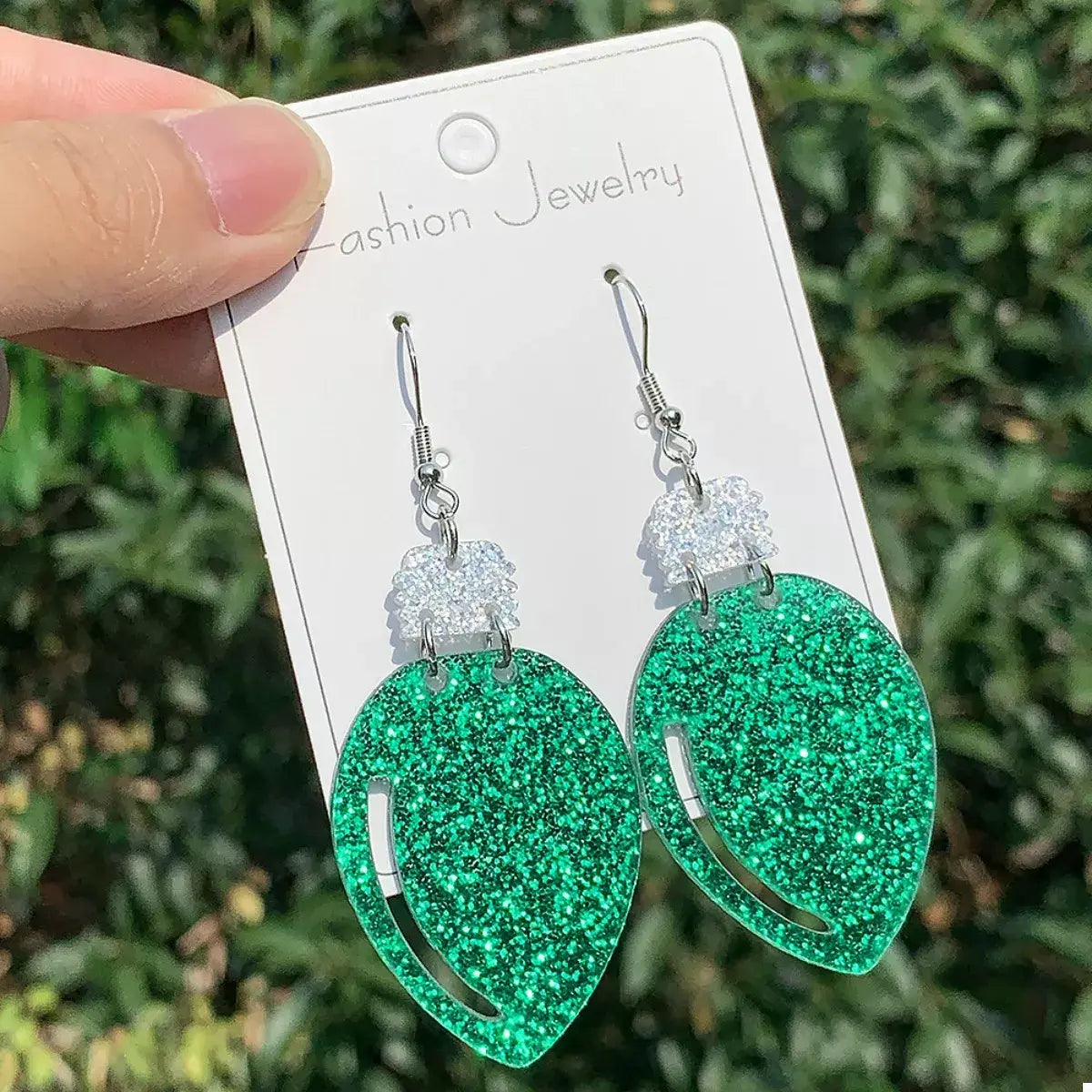 Green glittery earrings on a 'Fashion Jewelry' card held by a hand with a green leafy background.