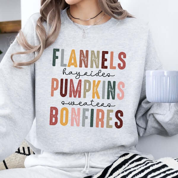 Gray sweatshirt with colorful text 'Flannels hayrides pumpkins sweaters bonfires' held by a person.