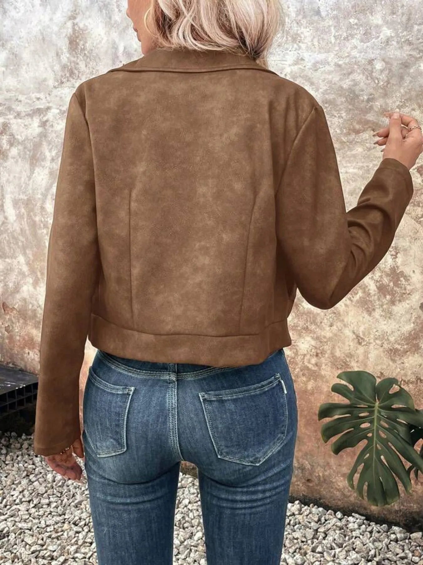 Person wearing a brown suede jacket and blue jeans against a textured wall.