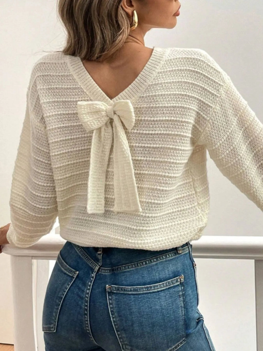 Woman wearing a cream sweater with a bow detail and blue jeans against a white background