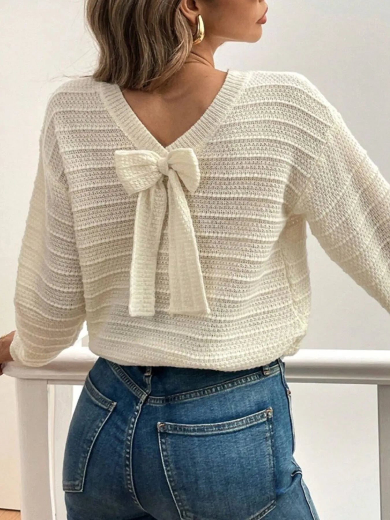 Woman wearing a cream sweater with a bow detail and blue jeans against a white background