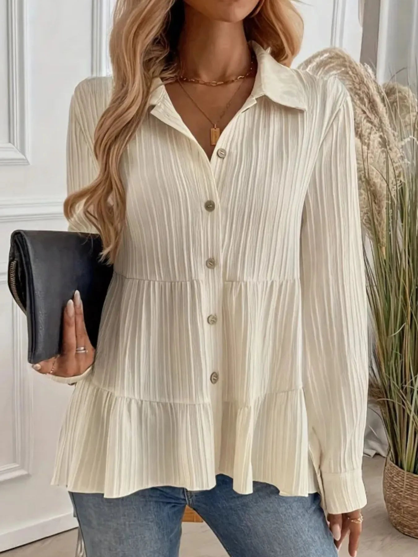Woman wearing a beige pleated blouse holding a black clutch in a neutral setting.