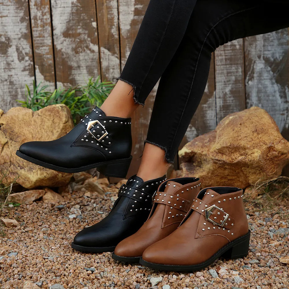 Black and brown studded ankle boots worn by a person standing on a rocky ground with a wooden fence background.
