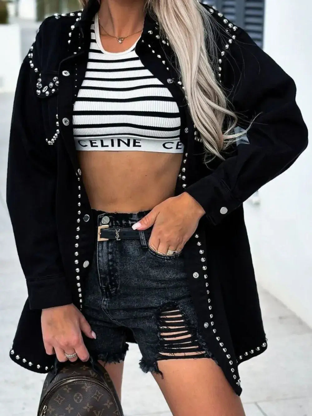 Person wearing a black studded jacket, striped crop top, and ripped shorts with a blurred background.