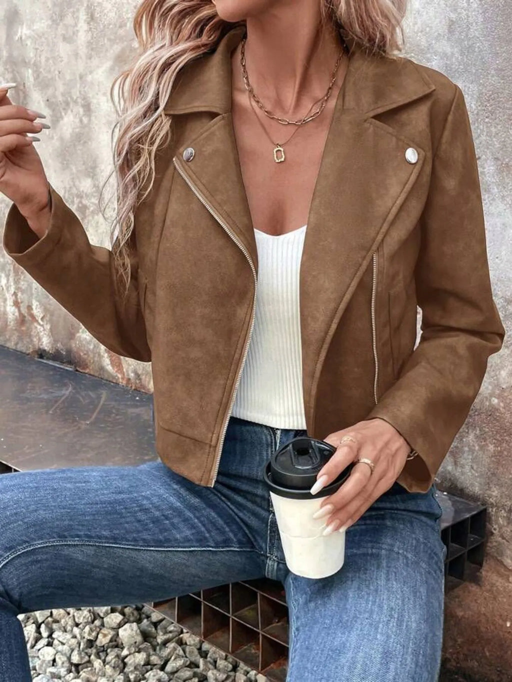 Woman wearing a brown suede jacket, white top, and blue jeans, holding a coffee cup.