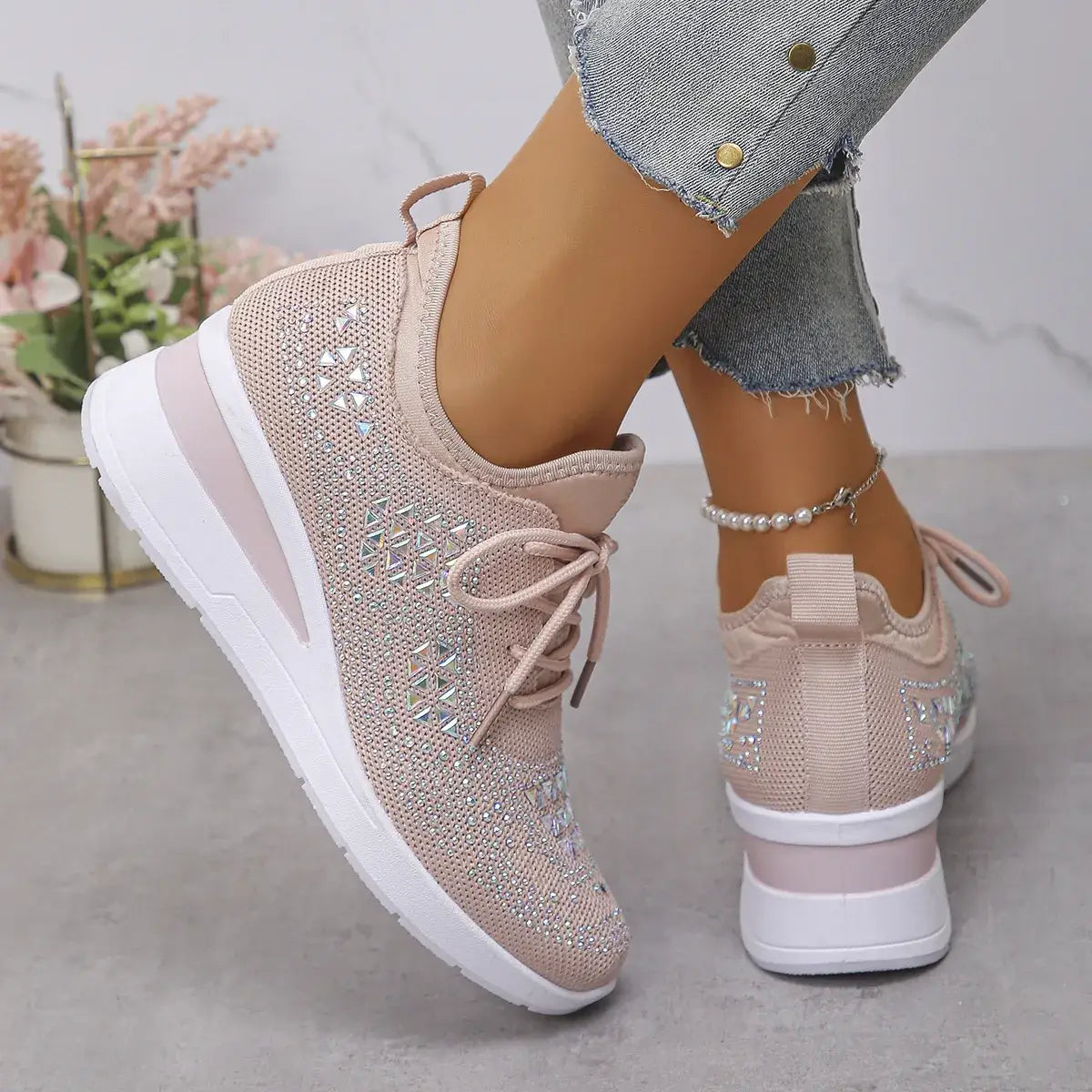 Beige sneakers with rhinestone details worn by a person, with a blurred background of flowers and a plant.