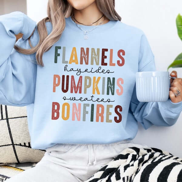 Person wearing a light blue sweatshirt with autumn-themed text, holding a mug.